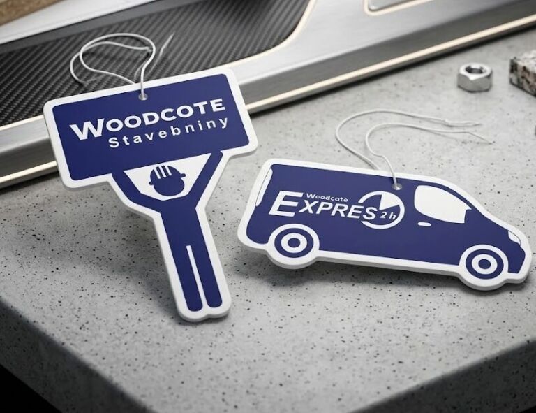 Dvě modré reklamní aromatické visačky pro Woodcote Stavebniny na šedém podkladu. Jedna ve tvaru postavy řemeslníka držícího ceduli a druhá ve tvaru dodávky s nápisem Woodcote Expres 2h